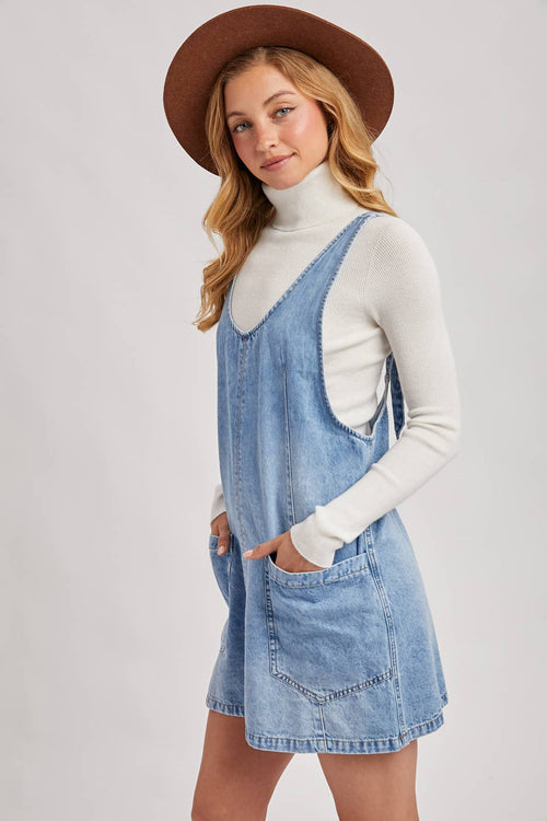 Woman wearing a denim dress over a white turtleneck with a brown hat on a light gray background