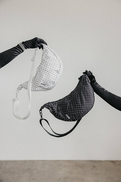 Two black and white polka dot crescent shoulder bags held up by black-gloved hands against a minimalist light gray wall.