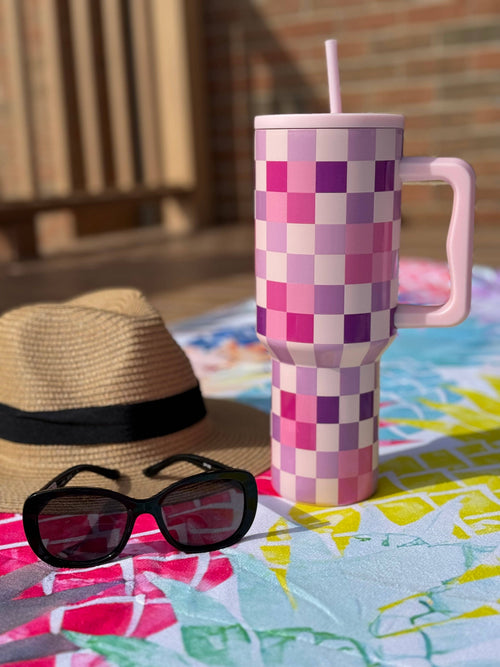 Checkered mug with a straw, sunglasses, and a hat on a colorful towel outdoors.
