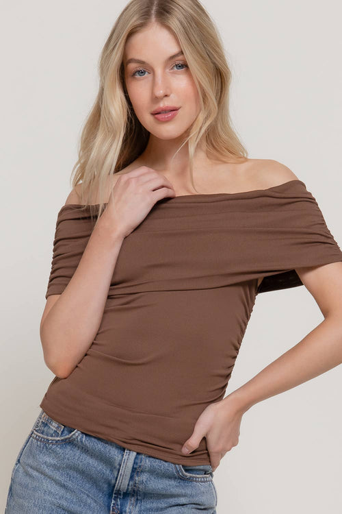 A woman modeling a brown off-the-shoulder short-sleeve top with ruched detailing, paired with blue jeans, against a plain light gray background.