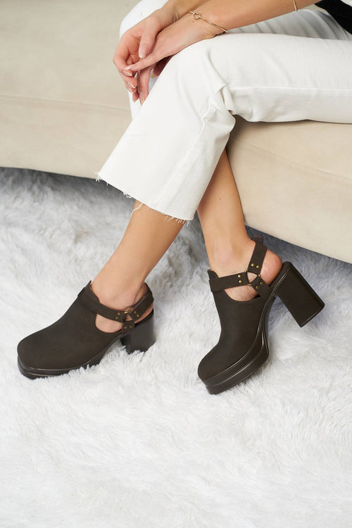 A person seated on a beige sofa wearing dark brown platform clogs with cutout straps and block heels, paired with white cropped pants on a white shag rug.