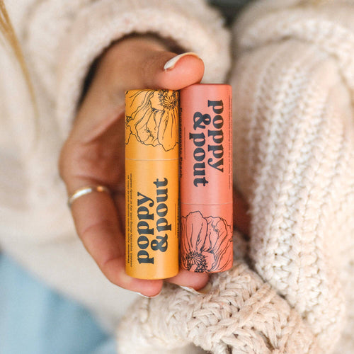 Two lip balm tubes from 'Poppy & Pout' held against a textured beige background.