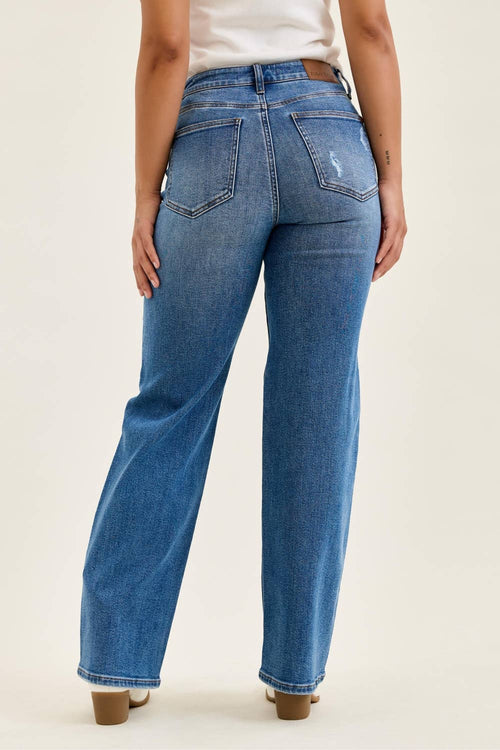 Rear view of a model wearing blue wide-leg jeans with a faded wash, paired with a white top and brown block-heel mules, against a plain off-white studio background.