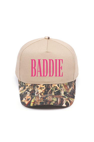 A beige baseball cap with 'BADDIE' printed in bright pink letters on the front, featuring a brown and green camouflage pattern visor.