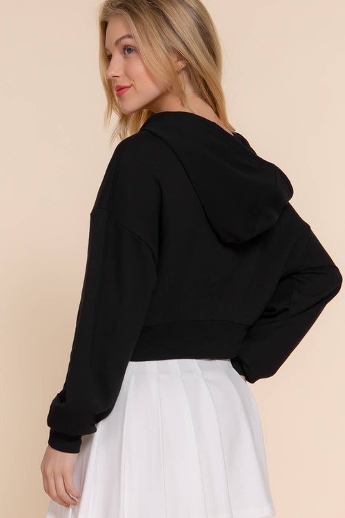 A woman shown from the back wearing a black cropped hoodie and a white pleated skirt, set against a solid light beige background.