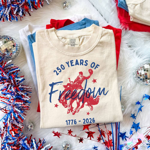 Beige t-shirt with '250 Years of Freedom 1776-2026' design on a festive background with red, white, and blue decorations.