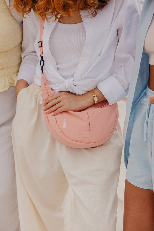 A close-up lifestyle shot of a person wearing a light pink crescent-shaped shoulder bag across their torso, styled with a white shirt and cream wide-leg pants.