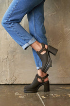A person wearing black platform clogs with ankle straps and block heels, paired with blue jeans, standing against a textured concrete wall.