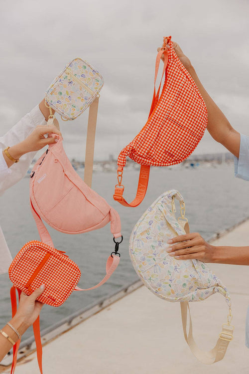 Four pastel sling bags in different colors and patterns are held up by multiple hands on a sunny day by the seaside. The bags include a coral orange gingham pattern, a soft pink solid color, a light peach solid color, and a cream floral print.