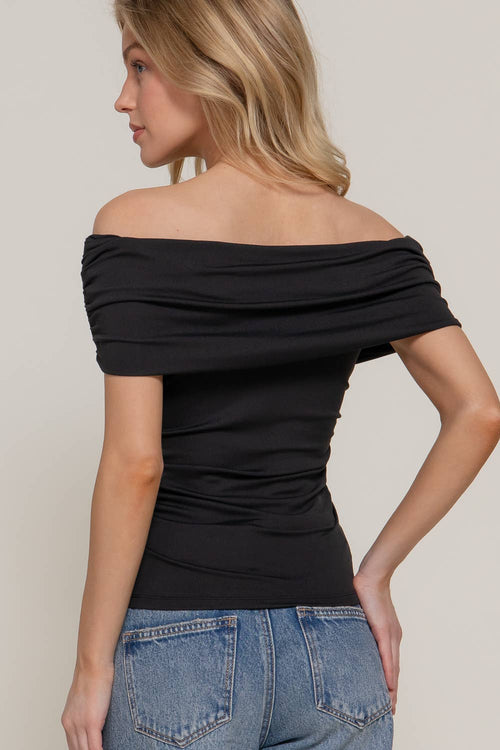 A woman wearing a black off-the-shoulder short-sleeve top and blue jeans, shown from a side-back angle against a plain light beige studio background.