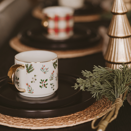 Decorative teacup with floral design on a black saucer, surrounded by greenery and a gold Christmas tree.