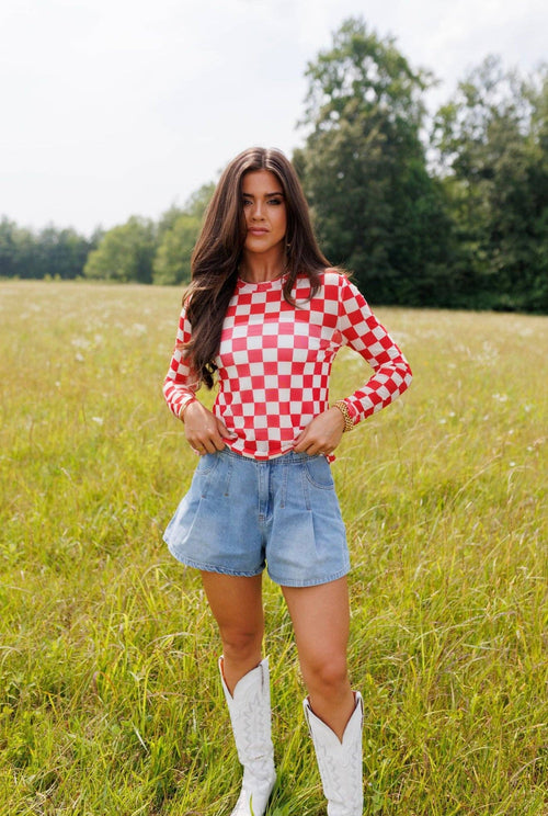 Woman in a red and white checkered shirt, denim shorts, and white boots standing in a grassy field.