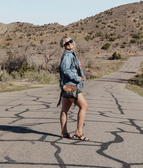 Woman in denim jacket and shorts standing on a desert road with mountains in the background