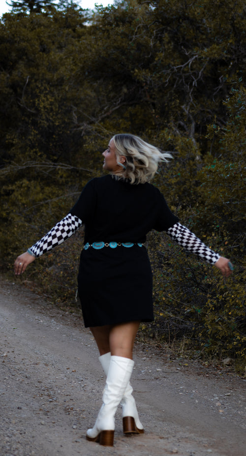 Woman in a black dress with checkered sleeves and white boots standing on a path with trees in the background