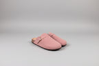 A pair of dusty pink suede clogs with a single strap and a gold-tone buckle, resting on a dark gray surface against a light gray background.