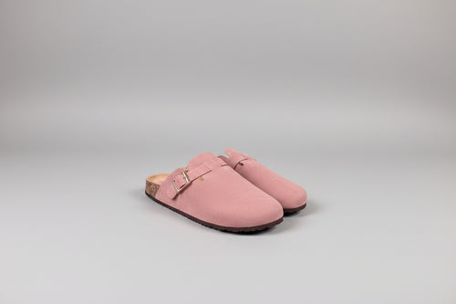 A pair of dusty pink suede clogs with a single strap and a gold-tone buckle, resting on a dark gray surface against a light gray background.