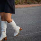 Person wearing a black dress with a graphic design, white knee-high boots, and checkered sleeves outdoors.