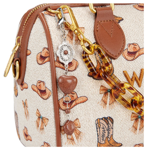Close-up of a handbag featuring a cream canvas body with a brown cowboy hat and bow print, brown leather trim, gold-tone hardware, and a dangling charm with a heart and silver medallion.