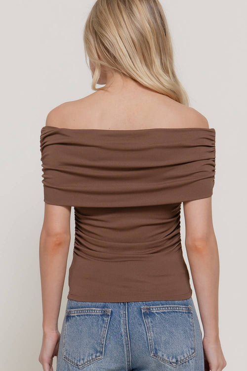A woman shown from the back wearing a brown off-the-shoulder ruched top and blue jeans against a light gray studio background.