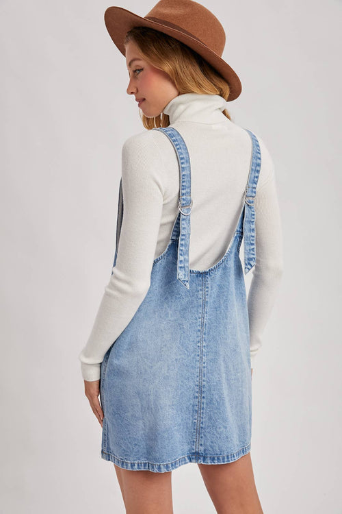 Woman wearing a denim pinafore dress over a white long-sleeve shirt with a brown hat on a light gray background