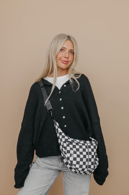 Woman wearing a black cardigan and light gray pants with a checkered bag on a beige background