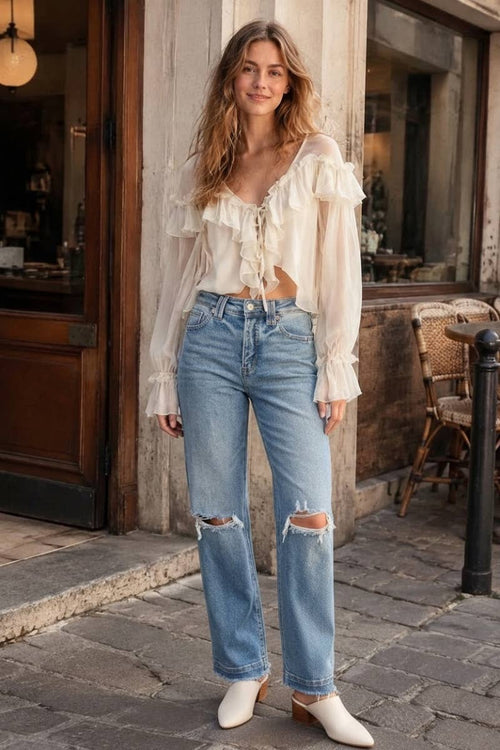 A woman stands on a cobblestone street in front of a rustic shop, wearing a cream ruffled long-sleeve crop top, light-wash distressed blue jeans, and white pointed-toe mules.