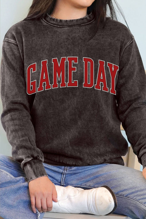 A person wearing a dark washed charcoal sweatshirt with 'GAME DAY' printed across the chest in red varsity letters with white outlines, paired with blue jeans and a white boot.