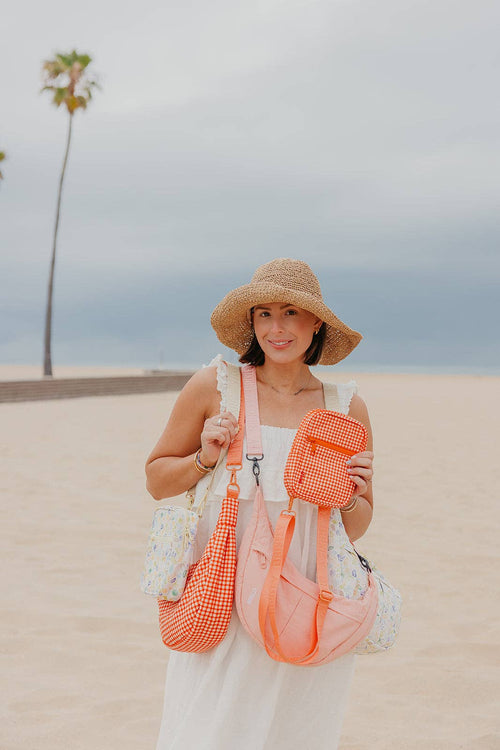 A woman on a sandy beach wearing a straw sun hat and white dress, carrying multiple orange and white patterned bags including shoulder bags, a crossbody, and a small pouch.