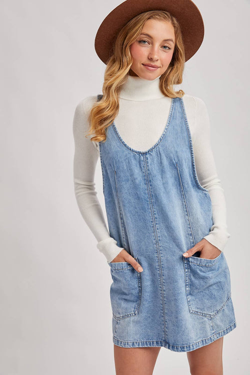 Woman wearing a denim dress over a white turtleneck with a brown hat on a light gray background