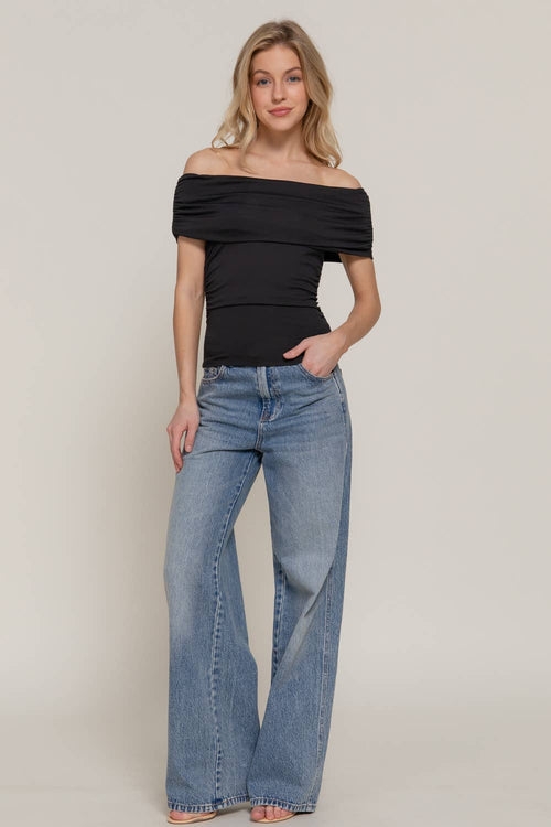 A woman modeling an off-the-shoulder black top and wide-leg blue jeans against a plain light beige studio background.