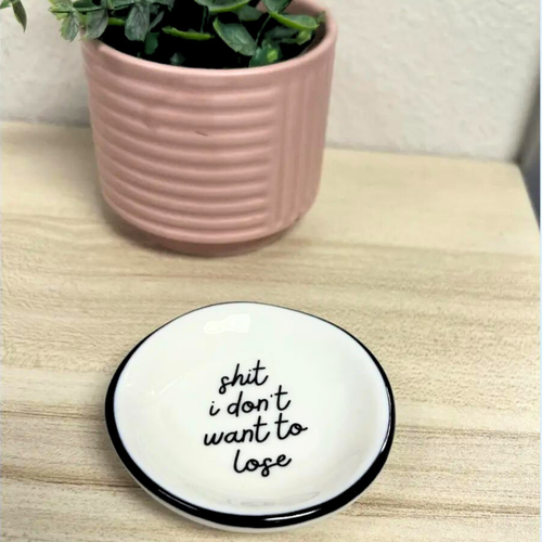 Round white plate with black rim and text 'shit i don't want to lose' on a wooden surface with a pink pot of plants.