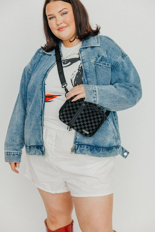 Person wearing a denim jacket, white shorts, and holding a black checkered bag against a plain background