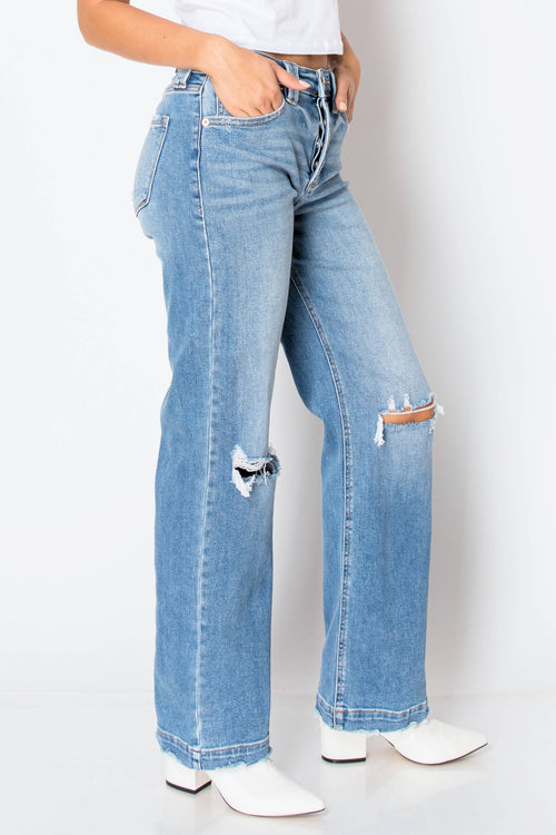 Light blue wide-leg denim jeans with distressed rips on the front of both legs, shown on a model wearing white pointed-toe heeled boots against a white studio background.
