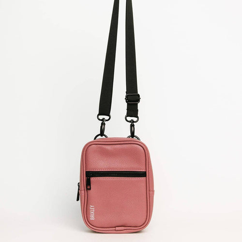 A pink pebbled faux-leather crossbody bag with a black adjustable shoulder strap, a black horizontal zippered pocket, and a small white 'BRAWLEY' logo on the bottom left corner, shown against a plain white background.