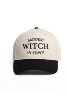 A two-tone baseball cap with an off-white crown and a black curved brim, featuring the text 'BADDEST WITCH IN TOWN' embroidered in black on the front.