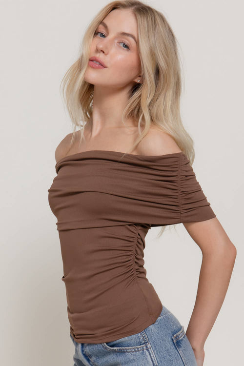 A woman wearing a ruched off-the-shoulder brown top and blue jeans, posing against a light gray studio background.