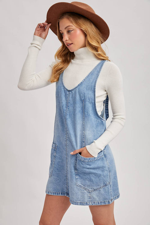 Woman wearing a denim pinafore dress with a white turtleneck and brown hat on a light gray background
